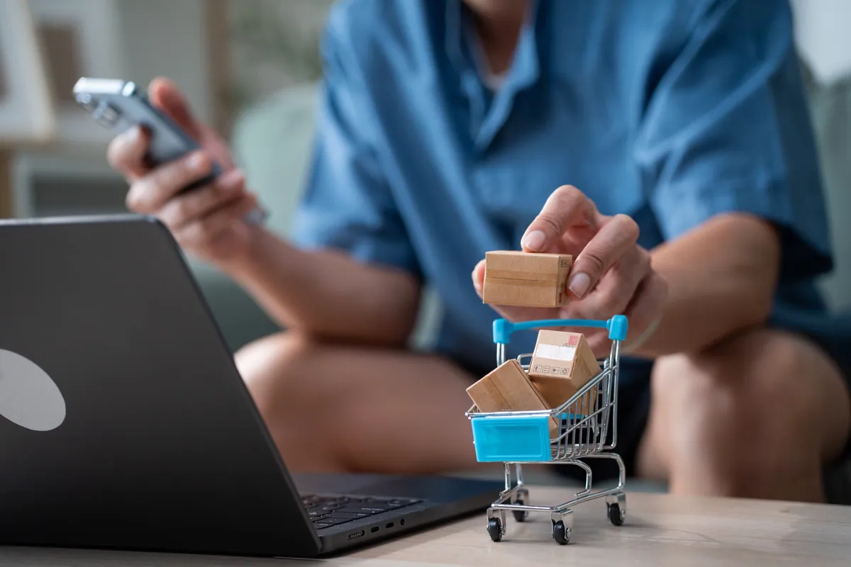 An online shopper with a phone, laptop, and miniature cart filled with parcels